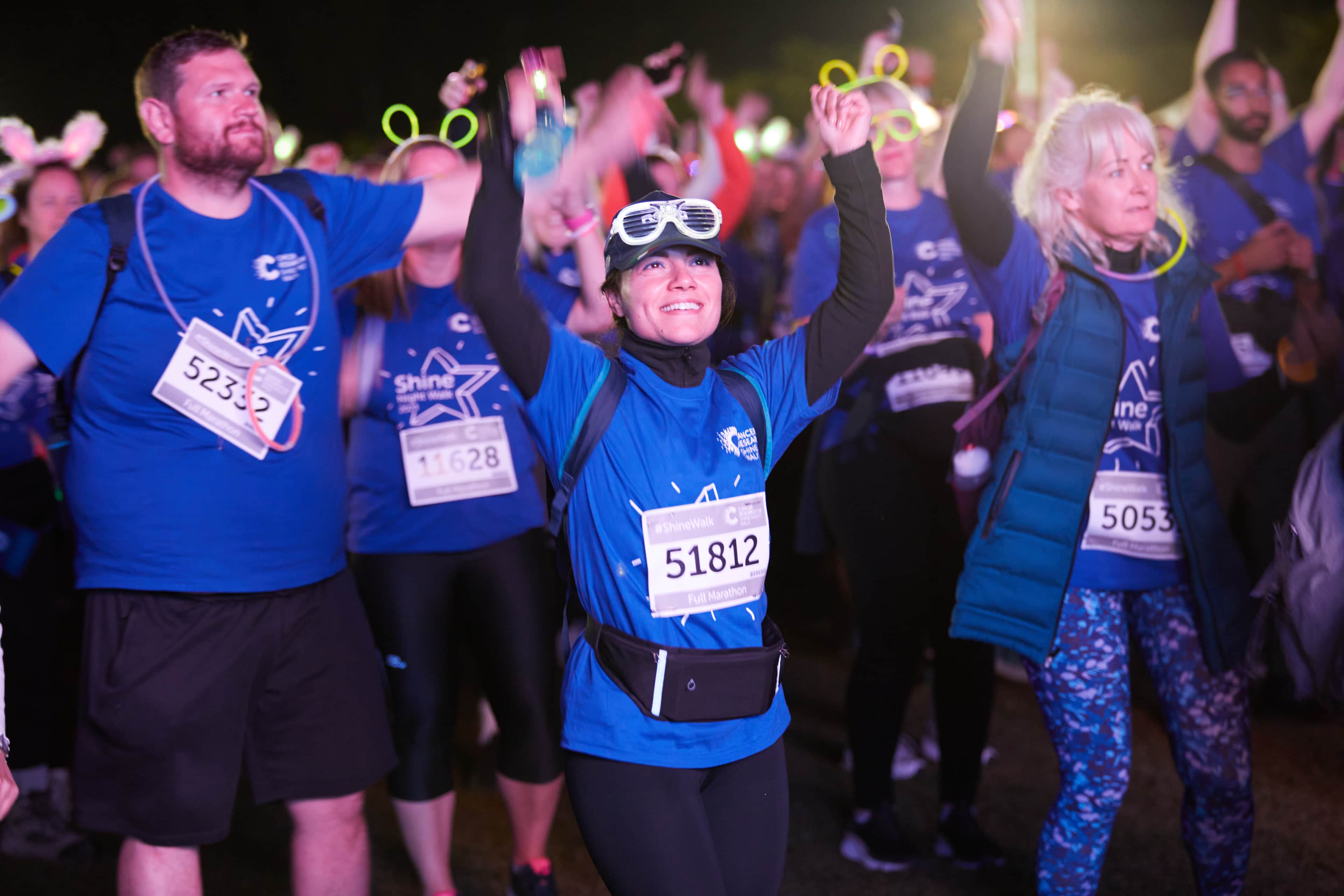 Shine night walk participant wearing white goggles surrounded by other participants celebrating with their hands in the air.