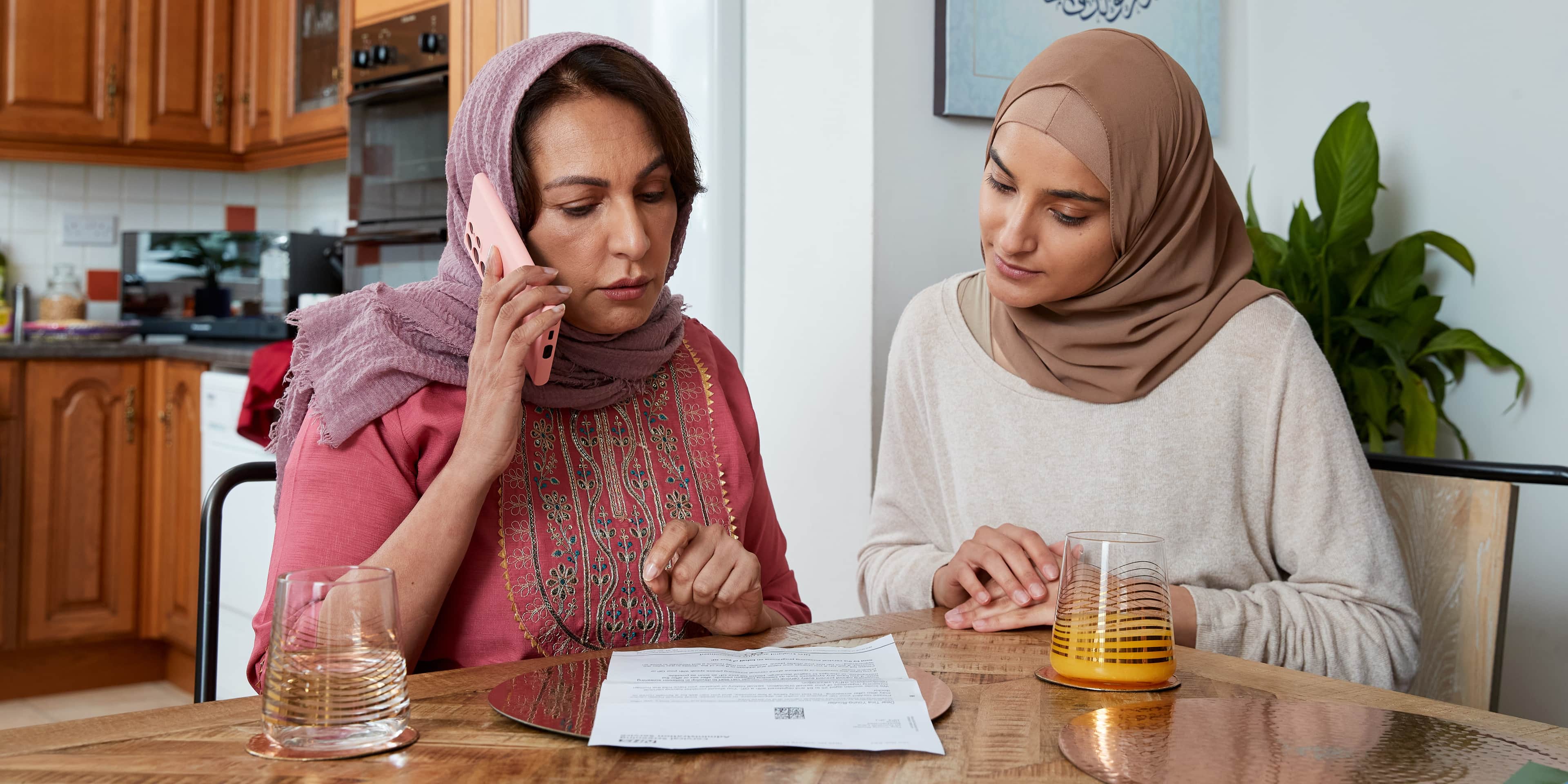 A mother booking a routine cervical or breast screening appointment on the phone with help from her daughter in the kitchen.