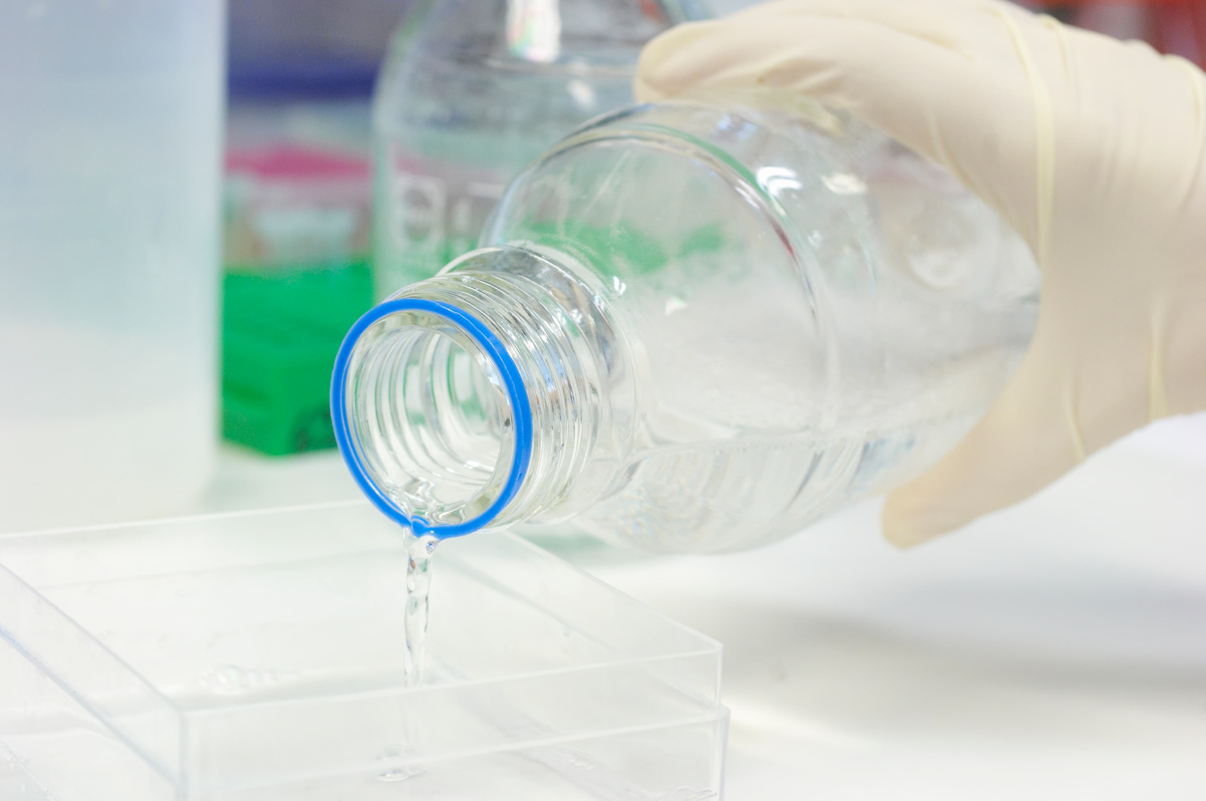 Clear liquid being poured from a Duran bottle into a square container in a lab.