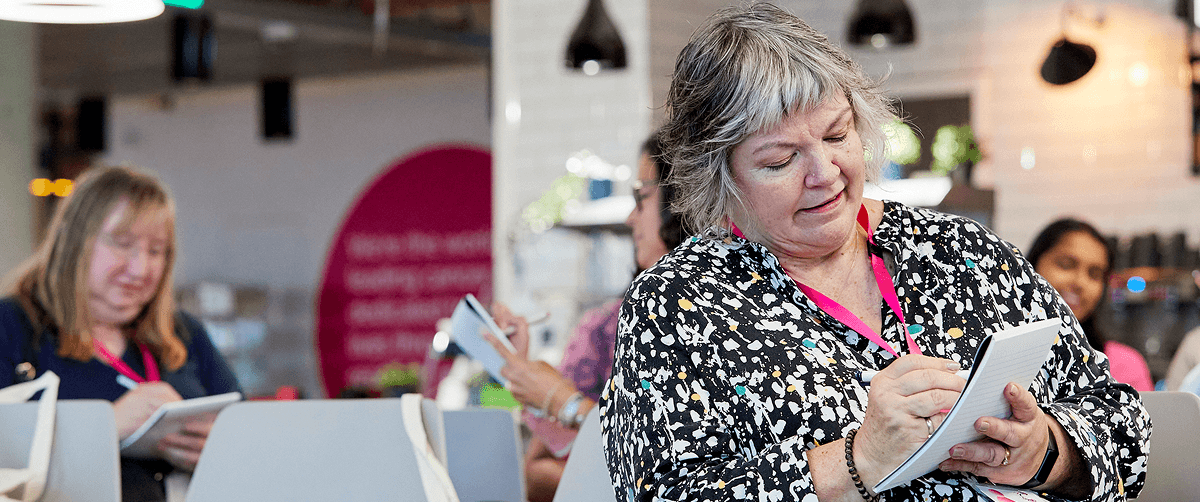 An image of a woman sat in a room that appears to be set up for an event. She is writing in a notebook.
