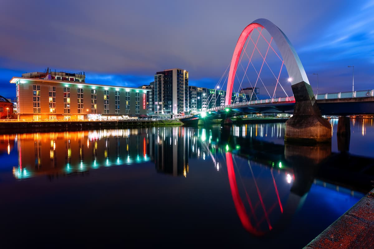 A night time view of the Clyde Arc and buildings lit up and reflecting in the River Clyde in Glasgow.