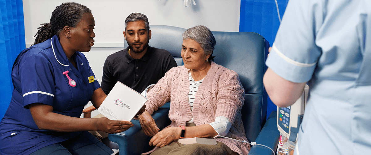 A nurse with a patient and their family talking them through some information with Cancer Research UK branding on.