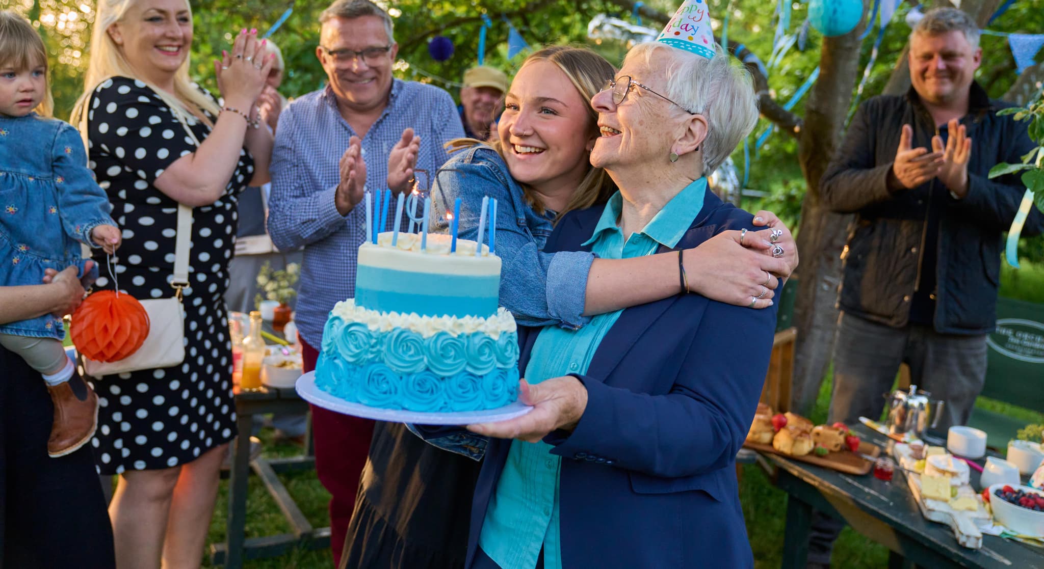 A woman celebrates her 75th birthday surrounded by friends and family. She is being hugged by a young woman.