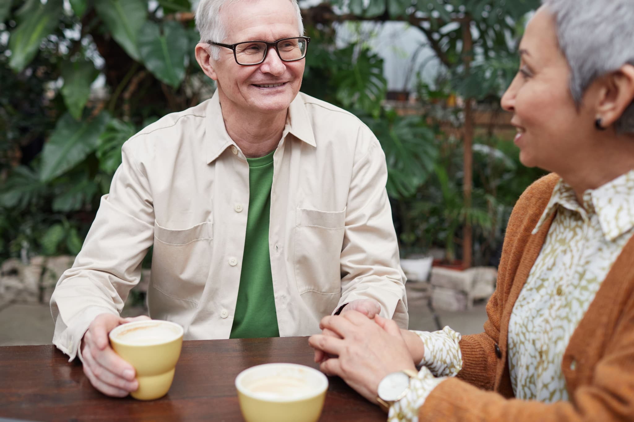 A man and are sitting at an outside table drinking coffee, the man is smiling to the woman.