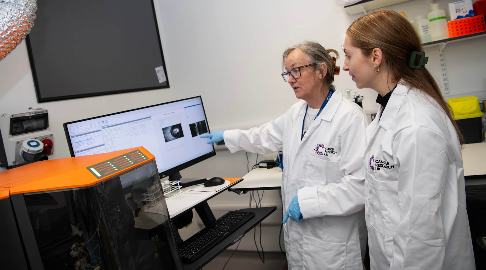 Image of two female researchers, wearing labl coats and looking at a computer screen.