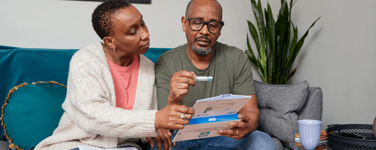 The photo shows a man with his partner on the sofa at home opening a bowel cancer screening test kit from NHS England.