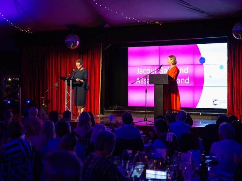 Two women standing on a stage at an Business Beats Cancer awards ceremony, one is presenting an award to the other.
